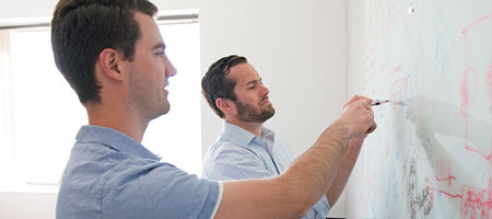Two businessmen talk while drawing on a whiteboard.
