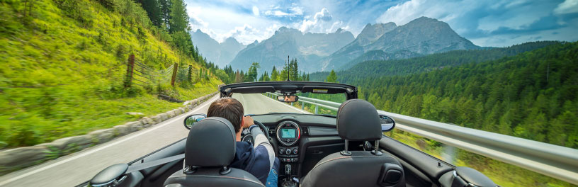 Backseat view of a convertible driving on a scenic mountain road, surrounded by forests and peaks under a cloudy sky.
