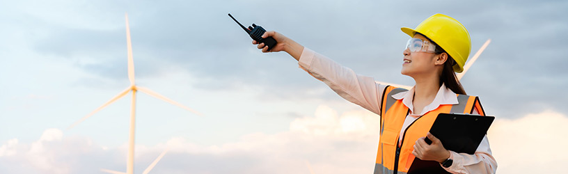 Female engineer wearing a safety helmet and reflective vest, holding a clipboard and using a walkie-talkie near wind turbines against a cloudy sky.