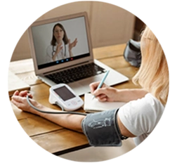 Person measuring blood pressure at home during a video call with a healthcare professional, taking notes.