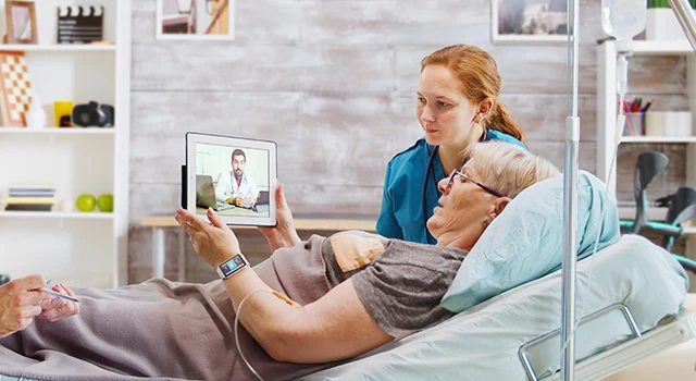 Elderly woman in home care video calls doctor, assisted by nurse in a calm, well-equipped room.