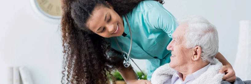 Healthcare worker smiling at elderly man in wheelchair, indoors with a warm, caring atmosphere.