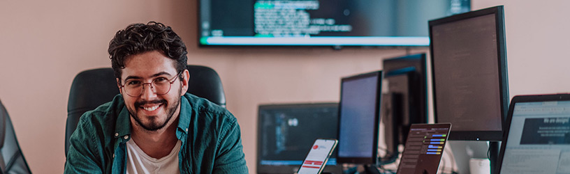 Smiling man in a casual shirt sitting at a desk surrounded by multiple computer screens and a smartphone in a modern workspace.