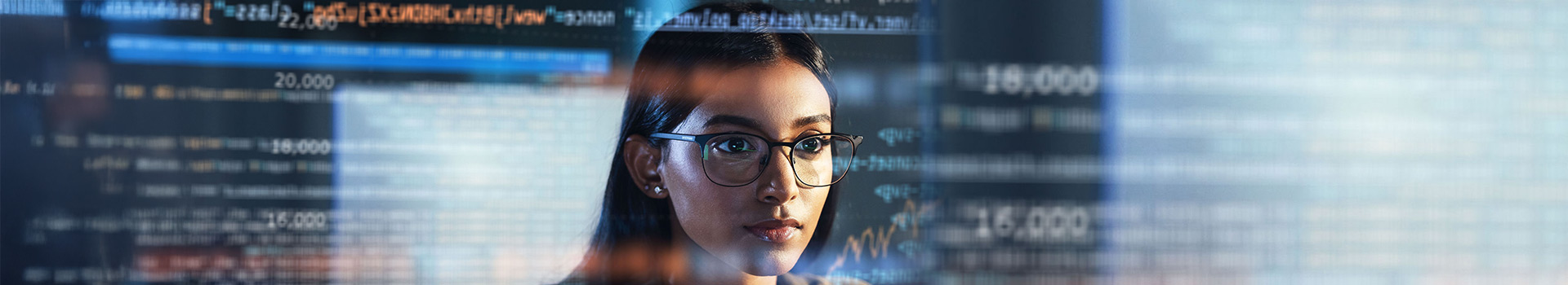 Focused woman wearing glasses analyzing data and code displayed on multiple digital screens in a technology environment.