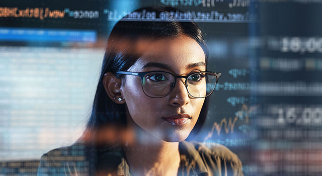 Focused woman wearing glasses analyzing data and code displayed on multiple digital screens in a technology environment.