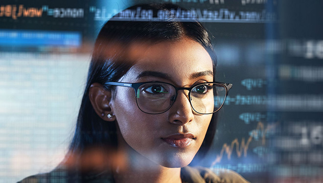 Focused woman wearing glasses analyzing data and code displayed on multiple digital screens in a technology environment.