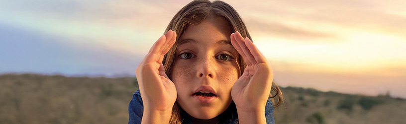 A young girl with freckles and brown hair gazes forward, framing her face with her hands as the sun sets over a scenic landscape in the background.